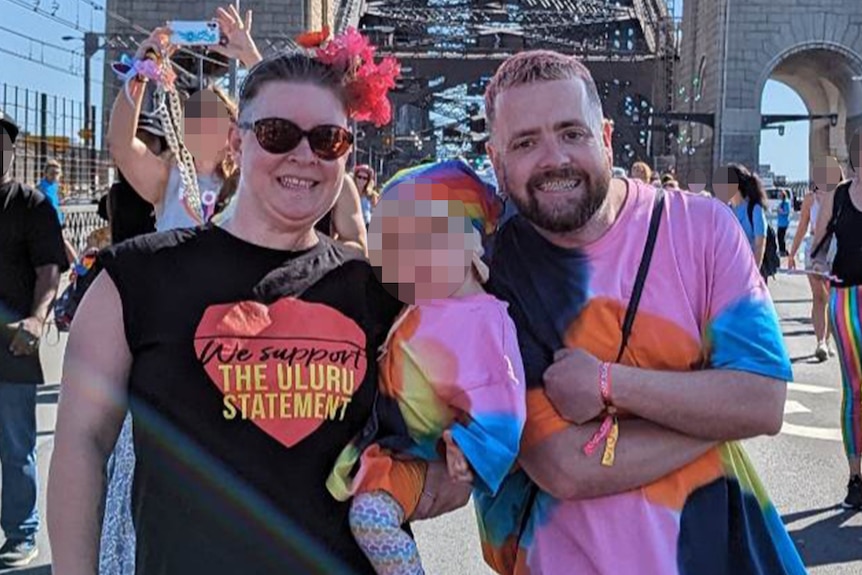 A man stands next to a woman holding a baby against the backdrop of the Harbour Bridge.