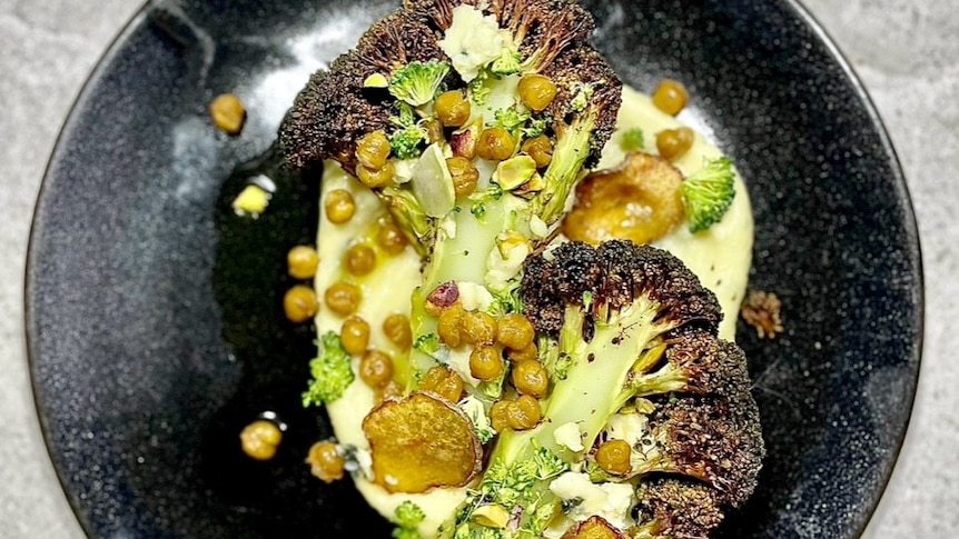 A black plate containing creamy potato mash, halves of charred broccoli, roasted chickpeas and pistatchios.