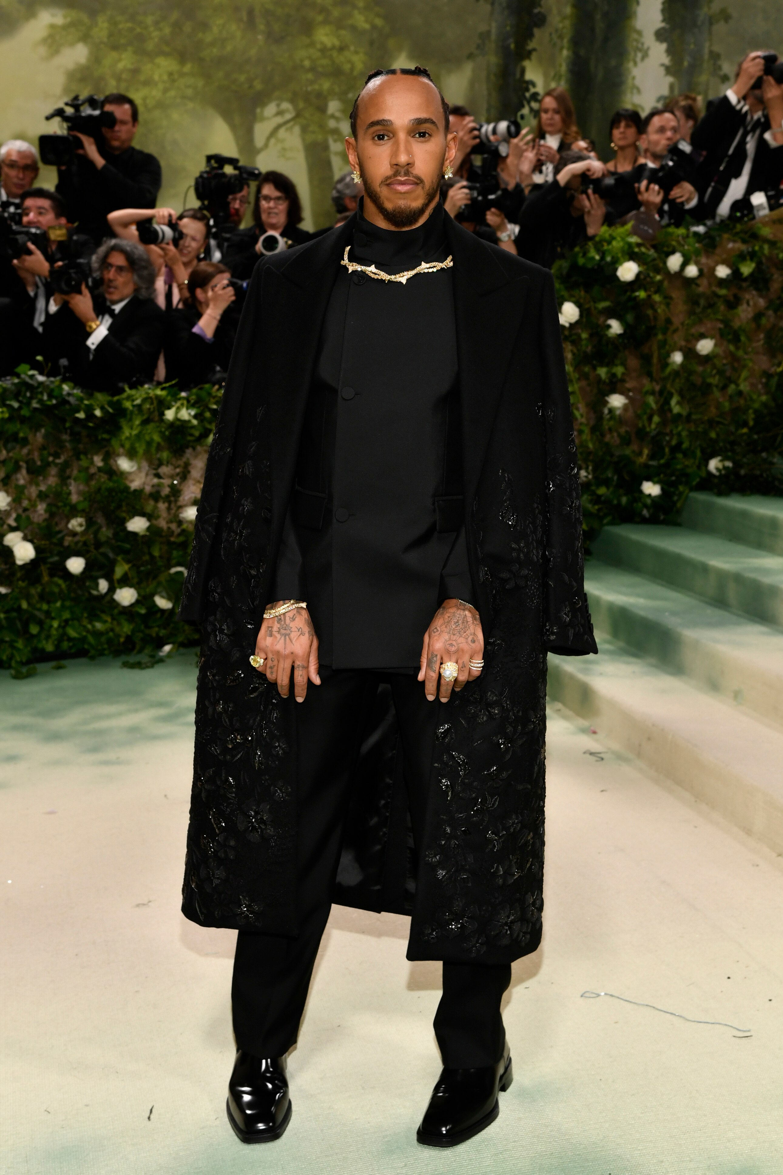 Lewis Hamilton wearing a black suit with a cloak clasped with a gold thorny necklace