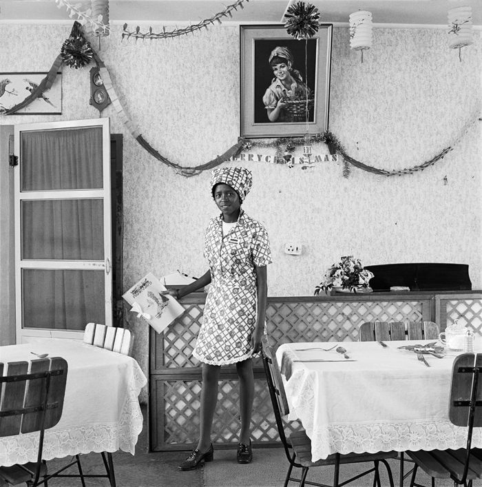 Young woman standing in diner, holding a menu in her hand and smiling. Black and white photo.