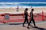 Two women in exercise gear walk past a sign blocking access to a beach. People can still be seen swimming