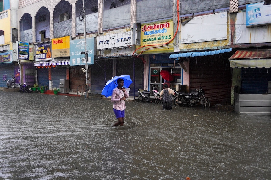 A man in pink shirt and blue shorts wades through a flooded street holding blue umbrella next to shops