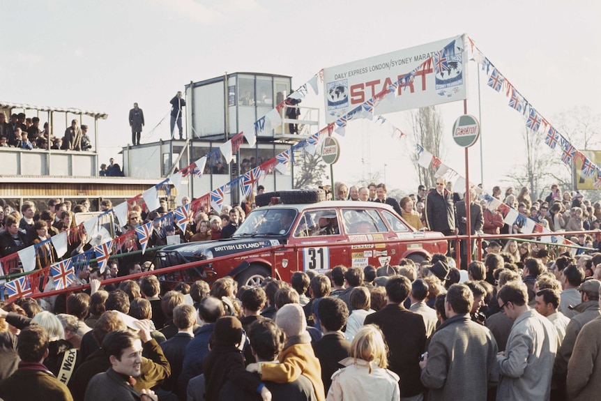 A red rally car sits at the start of a motor race surrounded by people.