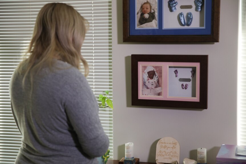 A young woman with blonde hair looks at picture frames showing baby photos and items hanging on a wall in a house.
