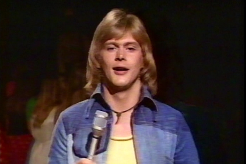 A man with long golden hair stands with a microphone in his hand.