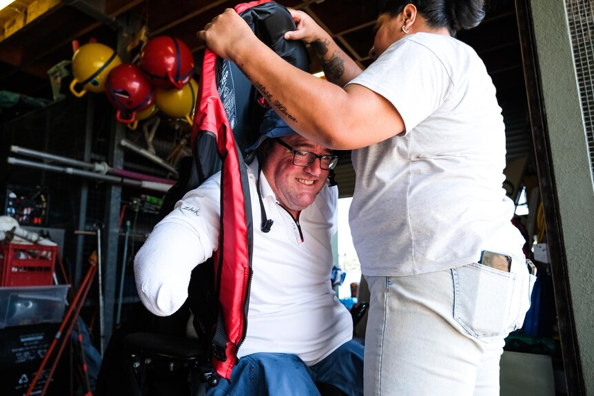 Man in bucket hat and black glasses and white shirt is being assisted to put lifejacket on. He has a visible arm amputation