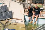 Two people sit on the side of a pool, surrounded by pieces of tin and solar panels from their destroyed accommodation