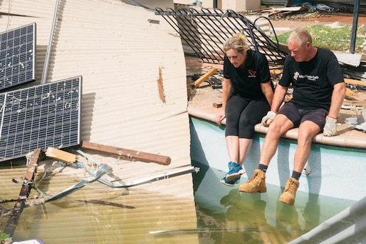 Two people sit on the side of a pool, surrounded by pieces of tin and solar panels from their destroyed accommodation