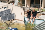 Two people sit on the side of a pool, surrounded by pieces of tin and solar panels from their destroyed accommodation