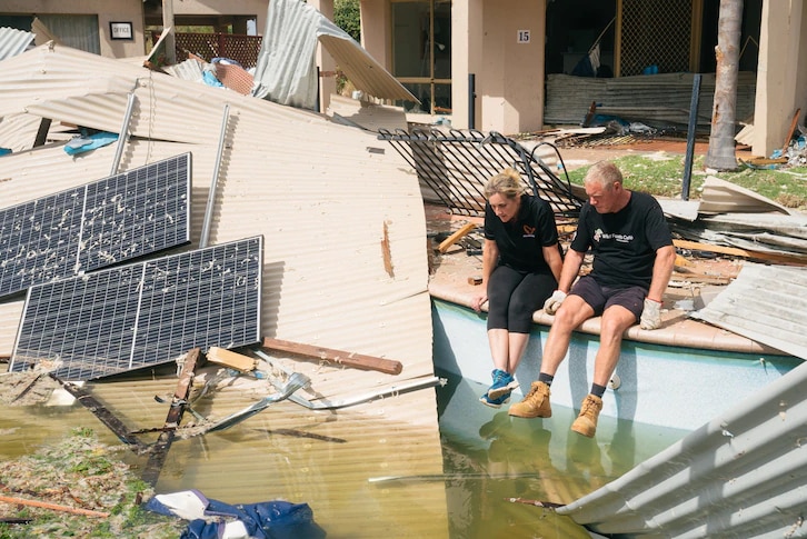 Two people sit on the side of a pool, surrounded by pieces of tin and solar panels from their destroyed accommodation
