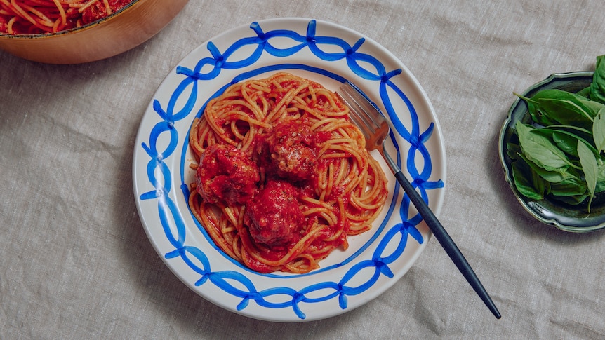 A pile of spaghetti and meatballs on a blue and white plate, with a fork and fresh basil in a bowl to the side