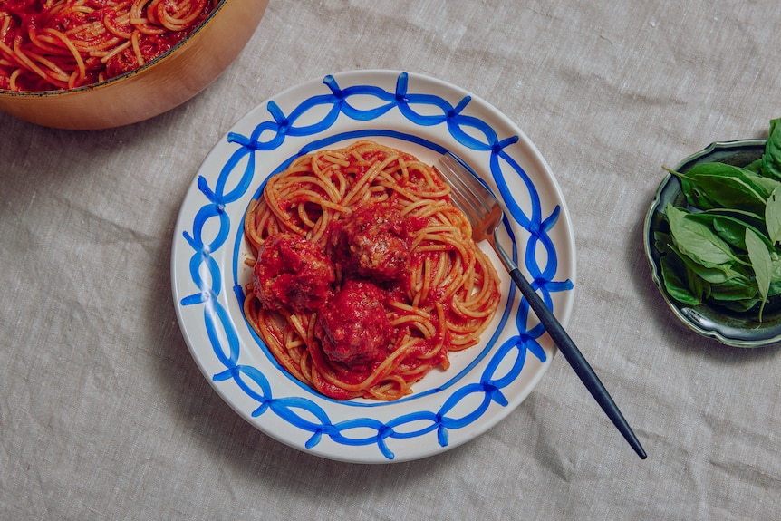 A pile of spaghetti and meatballs on a blue and white plate, with a fork and fresh basil in a bowl to the side