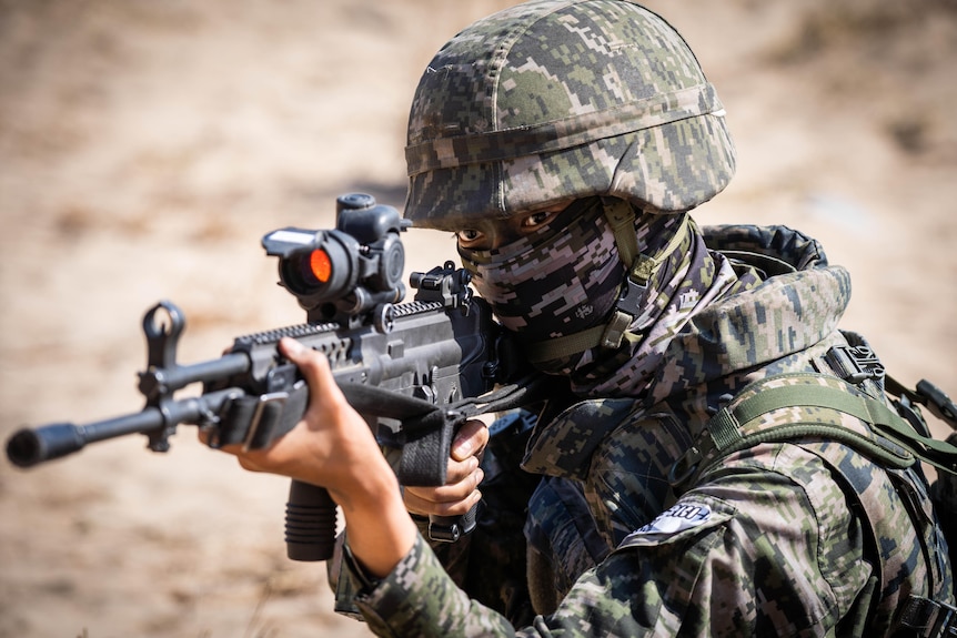 A person wearing full camouflage gear that covers up to just beneath their eyes holds an assault weapon