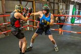 Two men wearing fitness wear and headgear sparring in a boxing ring with people watching and boxing equipment in the background 