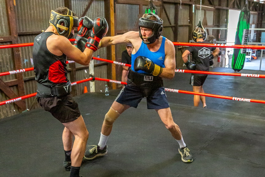 Two men wearing fitness wear and headgear sparring in a boxing ring with people watching and boxing equipment in the background 