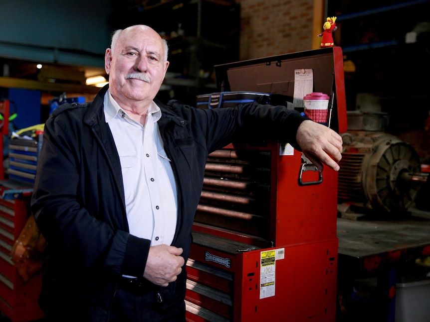 A balding man with white hair and a moustache leaning against a toolbox in a workshop wearing a white open collar t-shirt