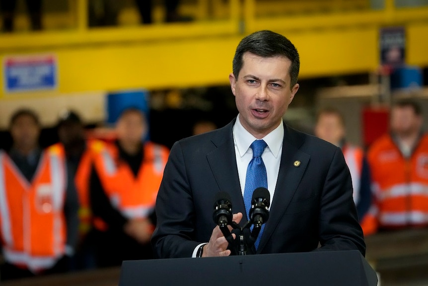 A young white man in a dark suit speaks in front of people wearing hi-vis uniforms at a construction site.