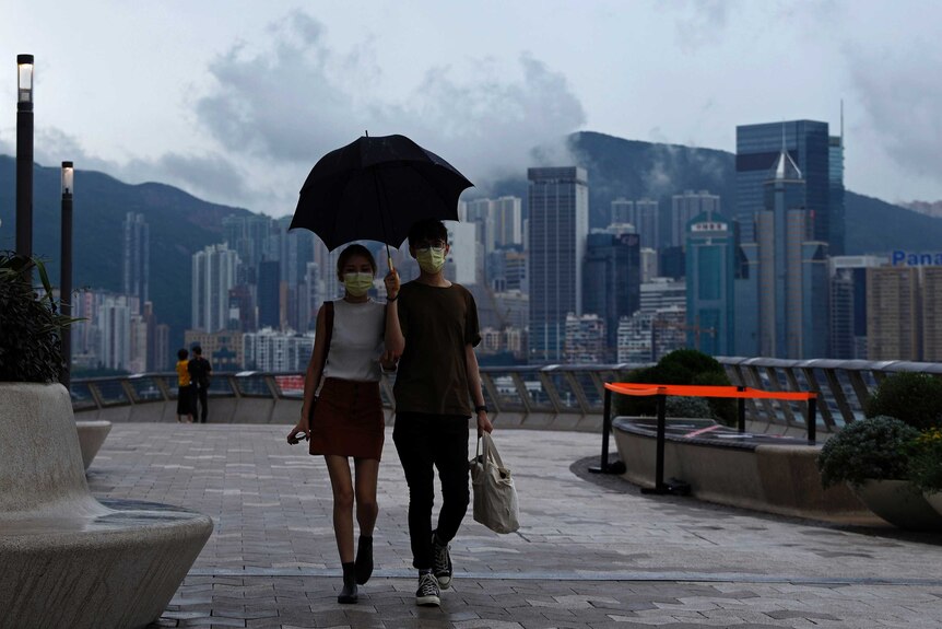 A man and woman walk under a black umbrella with the Hong Kong city visible in the background.