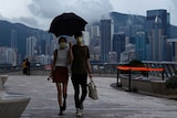 A man and woman walk under a black umbrella with the Hong Kong city visible in the background.
