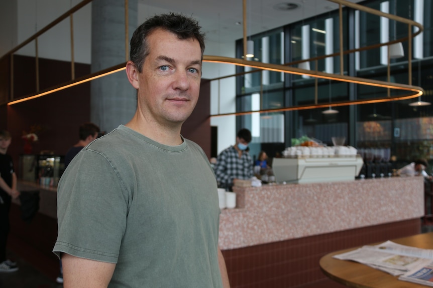 Tim smiles, standing in front of the counter of th cafe where a barista makes coffee.