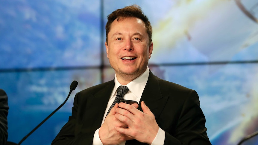 Elon Musk smiles while holding a phone to his chest, he is wearing a suit