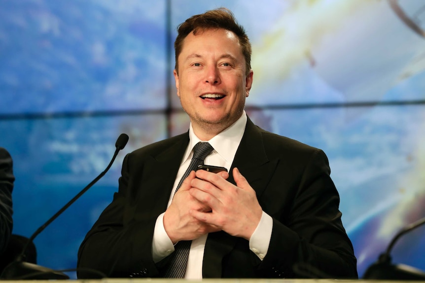 Elon Musk smiles while holding a phone to his chest, he is wearing a suit