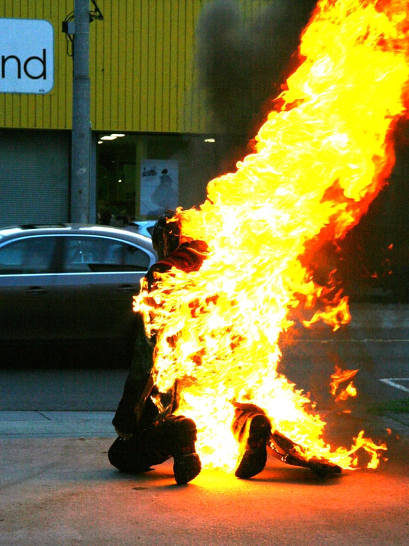 A man kneels on a city street set fully ablaze