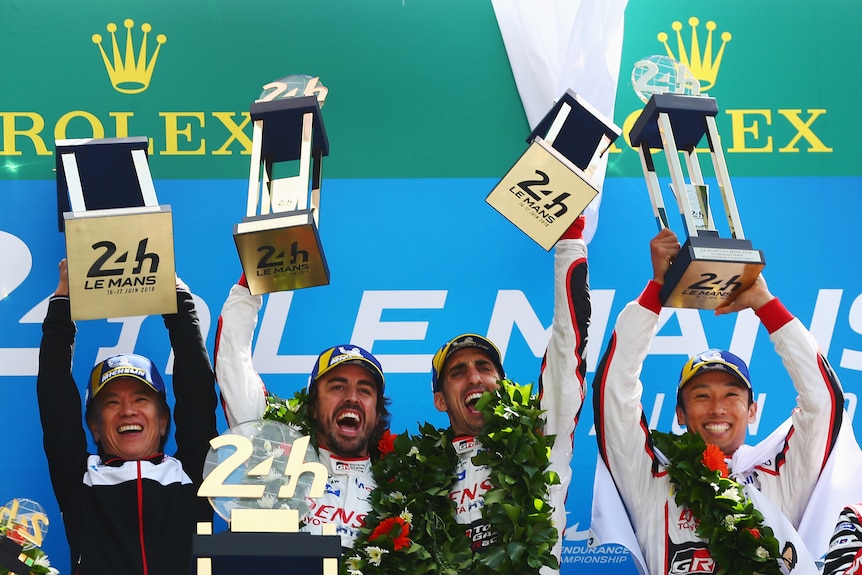 Four men stand on the top podium in celebration with trophies after winning the 24 Hours of Le Mans.