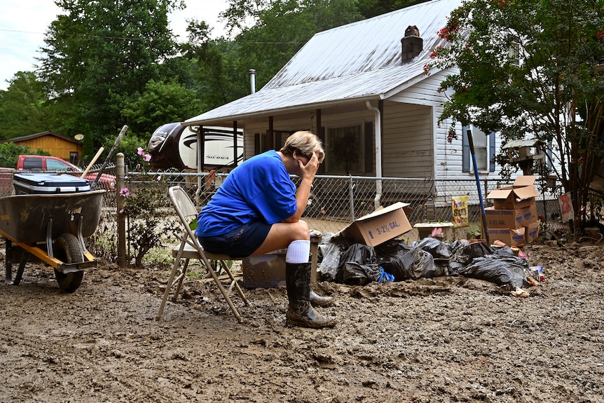 Woman in blue shirt and gumboots sits on chair with head in hands on her muddy lawn.