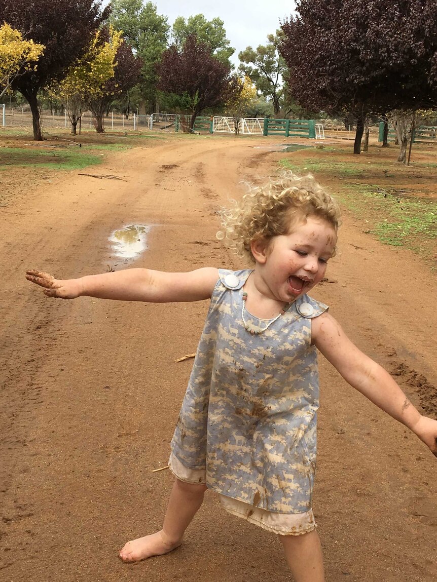 A small girl with mud on her face and hands frolics barefoot on a muddy property road.