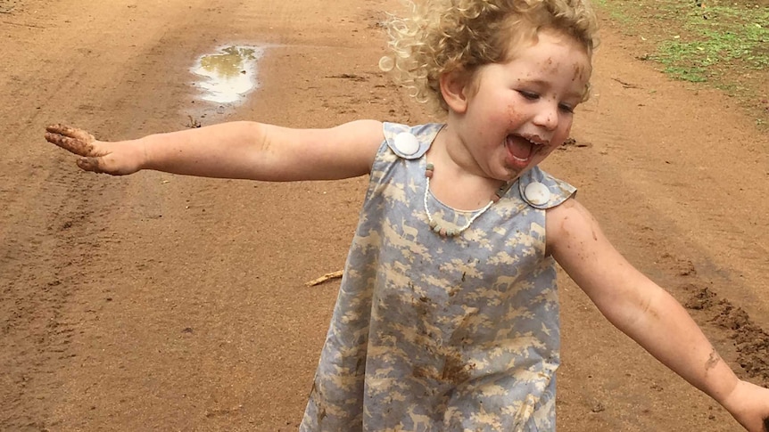 A small girl with mud on her face and hands frolics barefoot on a muddy property road.
