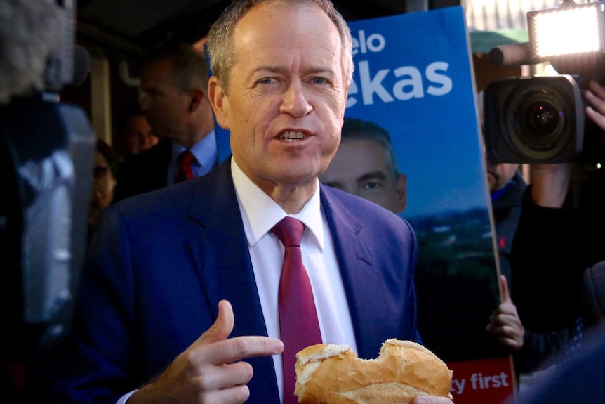 Bill Shorten points to a democracy sausage on bread with a bite incorrectly taken out of the side of the roll.