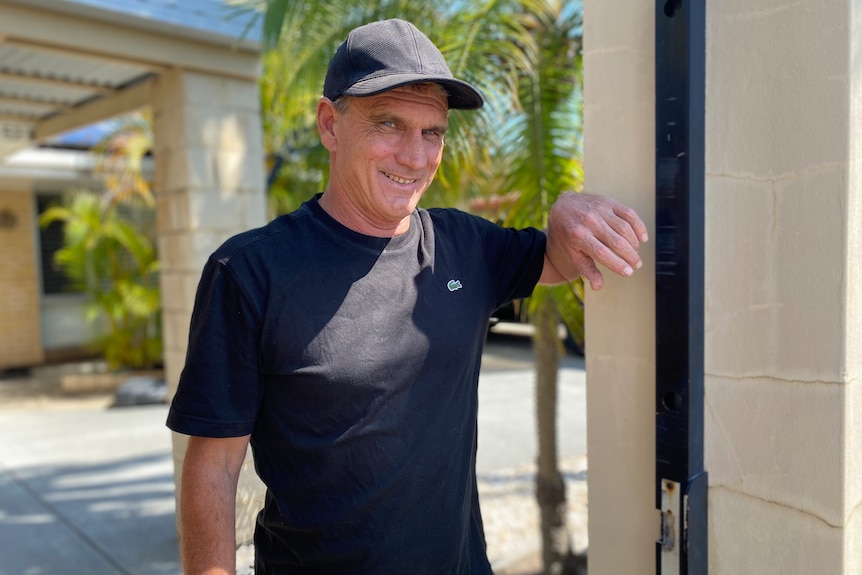 A man wearing a black t-shirt leans on his front gate.