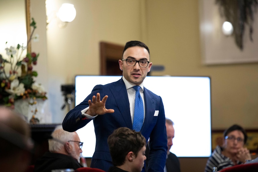 A young man wearing glasses and a blue suit gesticulates at a meeting