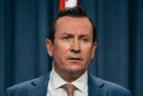 WA Premier Mark McGowan wearing a suit, standing at a lectern in front of a blue curtain and australian flags.