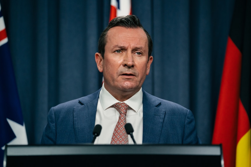 WA Premier Mark McGowan wearing a suit, standing at a lectern in front of a blue curtain and australian flags.