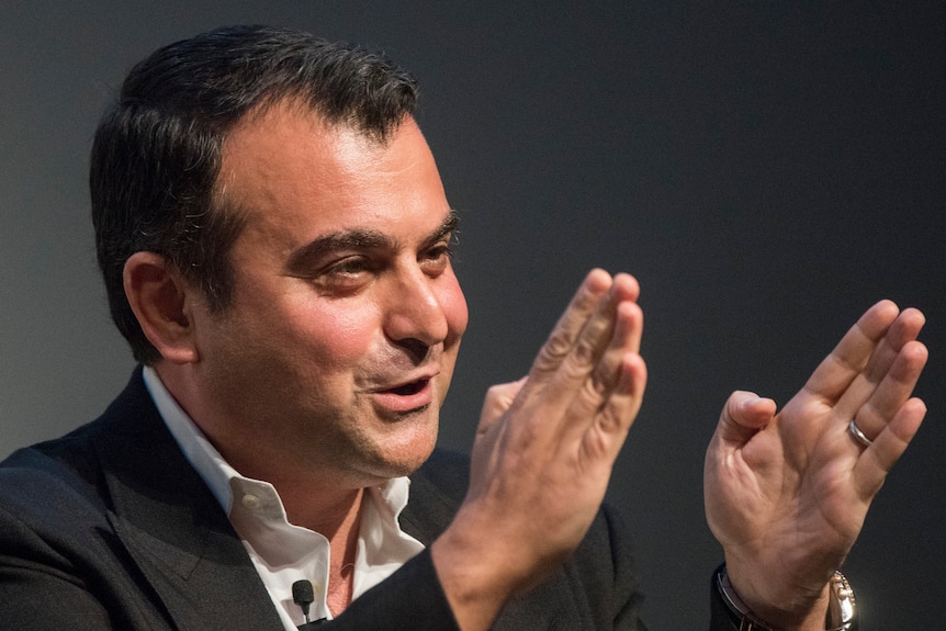 A man in a suit with dark hair gestures with his hands while speaking 