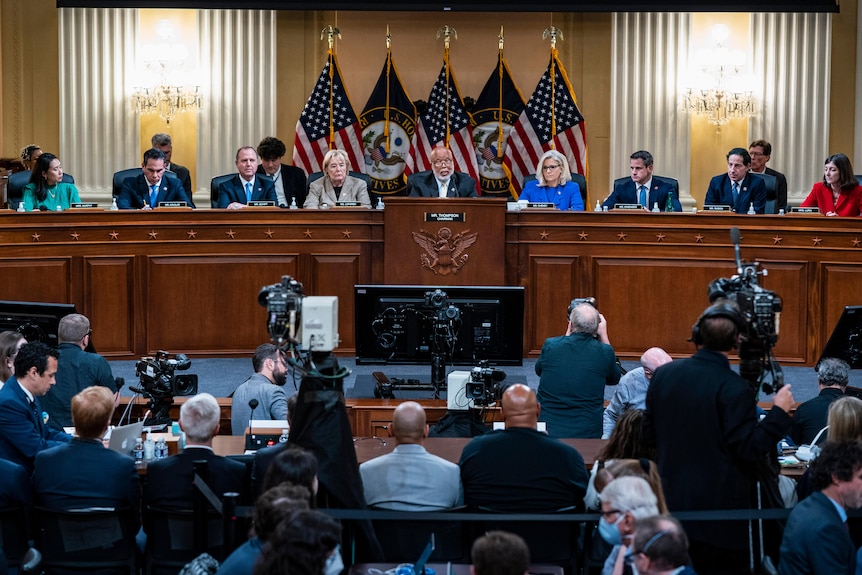 Professionally dressed people sit on a panel infront of US flags on display before the media and observers