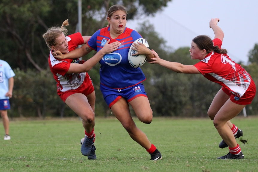 Caitlan Johnston, wearing the blue and red of the Newcastle Knights, bursts through two defenders with ball in hand.