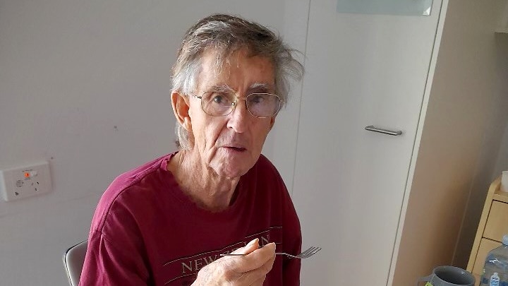 An elderly man with glasses sitting at a table lifting a fork with food to his mouth