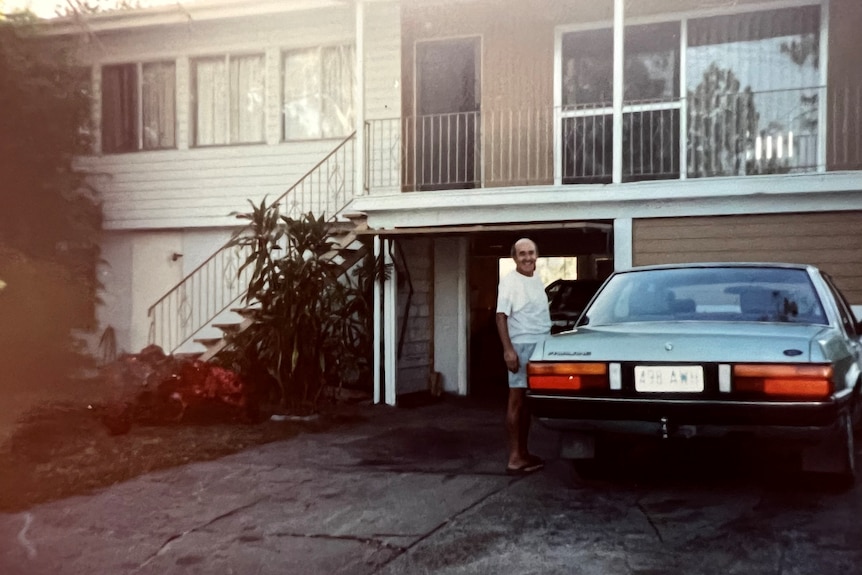A man stands in front of a house with a blue car. It's an old picture.