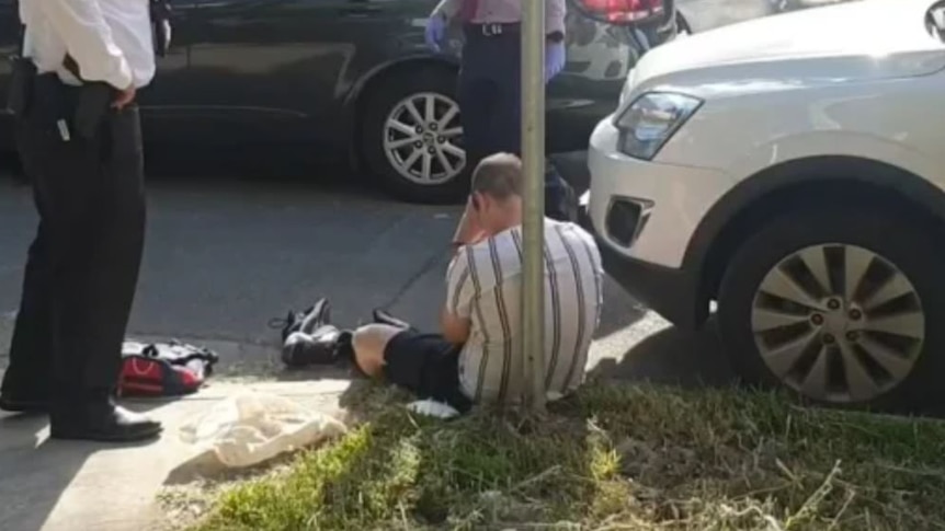 A man in a striped shirt and shorts sits on the kerb as two detectives, with faces blurred, stand nearby