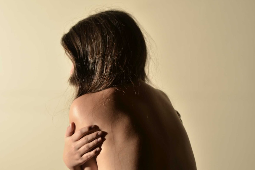 A woman sitting up in bed with no top on hugging herself tightly
