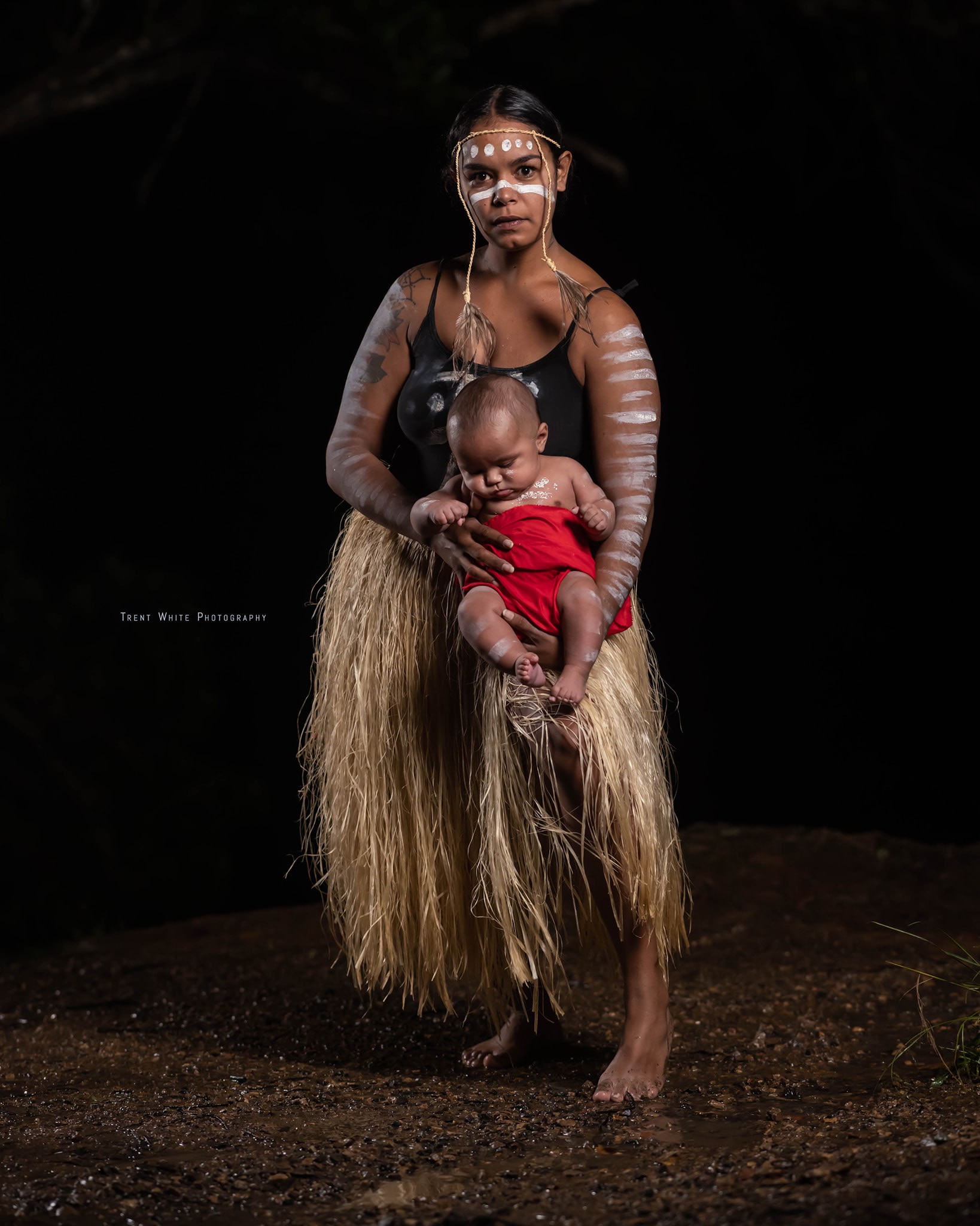 An Indigenous woman in a grass skirt, holding her son. Both wear traditional paint