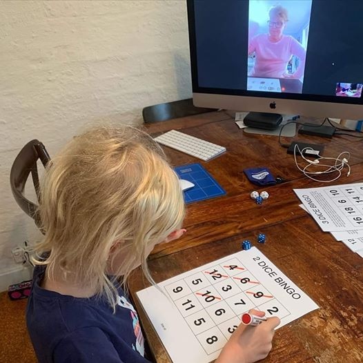 A child sits at a table with a bingo card, while a woman on a computer screen can also be seen playing