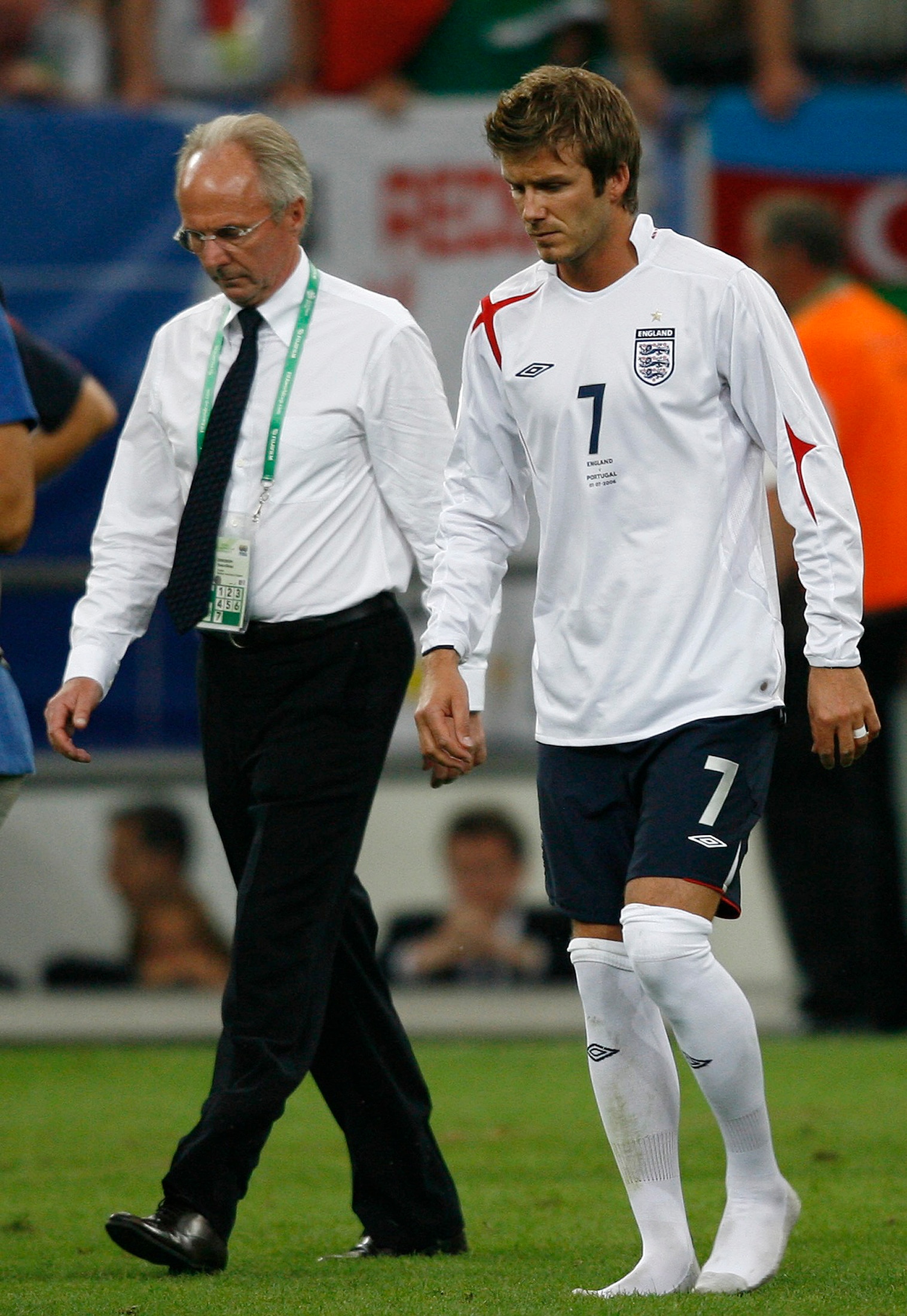 A middle-aged to older man in a business shirt and trousers walks beside David Beckham in football gear.