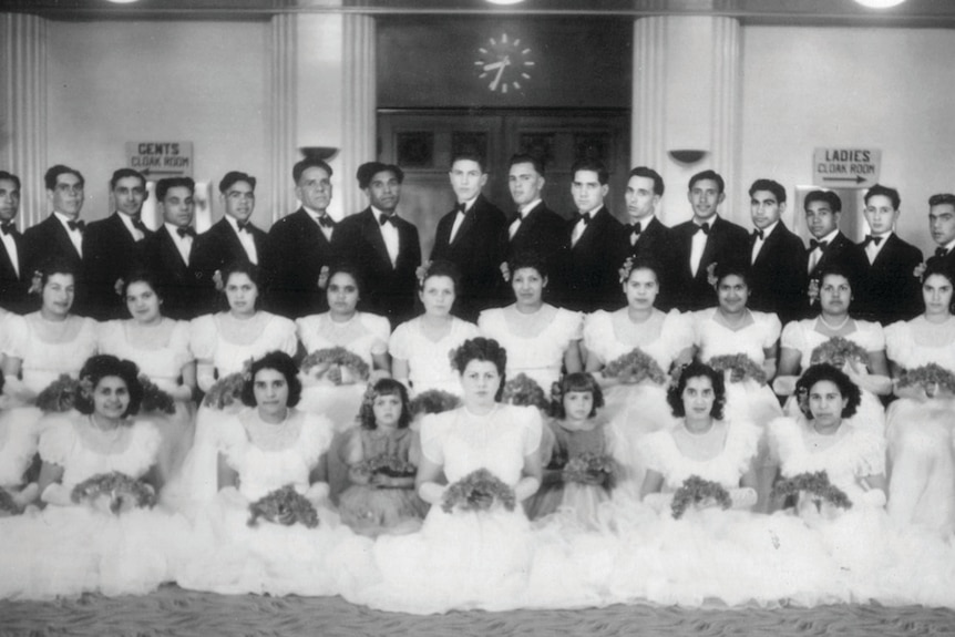 A black-and-white photograph of a group of 26 young Aboriginal women dressed in ball gowns. Suited men stand behind them.