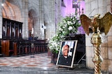 A picture of Archbishop Desmond Tutu stands on ground next to flowers in an archaic church