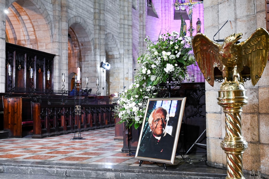 A picture of Archbishop Desmond Tutu stands on ground next to flowers in an archaic church