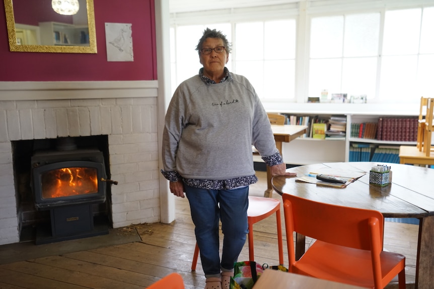 A woman in a grey jumper stands next to a fire and table.